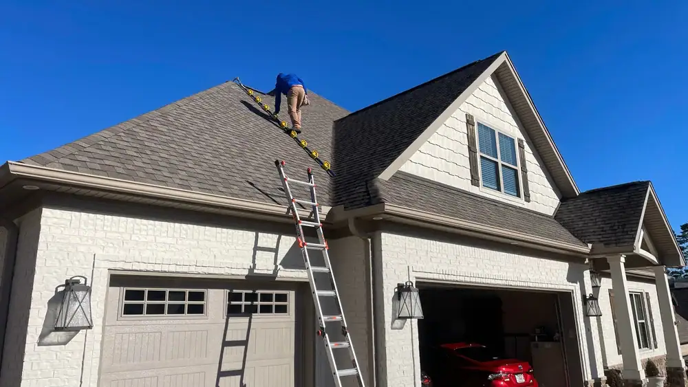 A person stands near the top of a steep house roof, holding onto a yellow ladder on the shingles. Another ladder leads up from the ground to the roof’s edge, next to a white garage—typical of home construction in Suffolk County, NY.