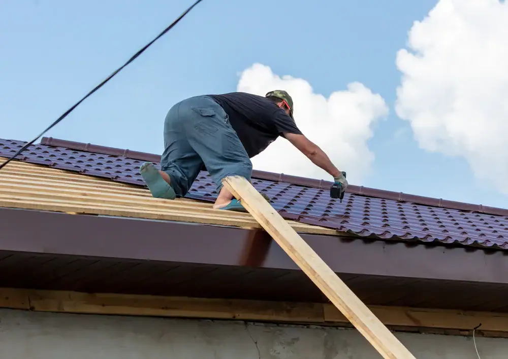 A person in work clothes and gloves kneels on a sloped surface, installing or repairing a metal roof—a common scene in home construction Suffolk County, NY. A wooden plank leads up to the roof under a blue sky with clouds in the background.