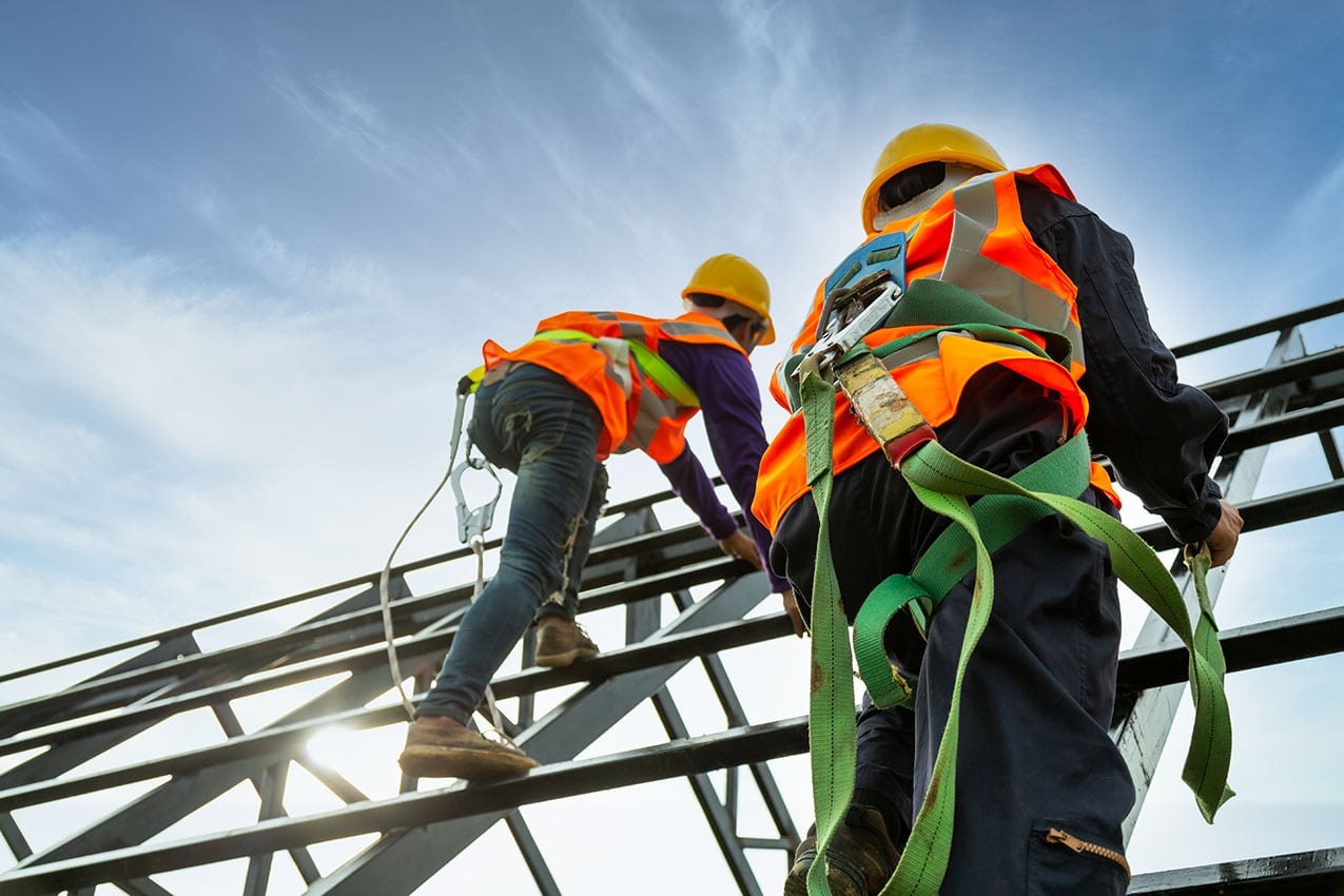 Two construction workers wearing safety harnesses, helmets, and reflective vests are climbing a steel structure against a blue sky, with the sun shining in the background—a common scene in home construction in Suffolk County, NY.