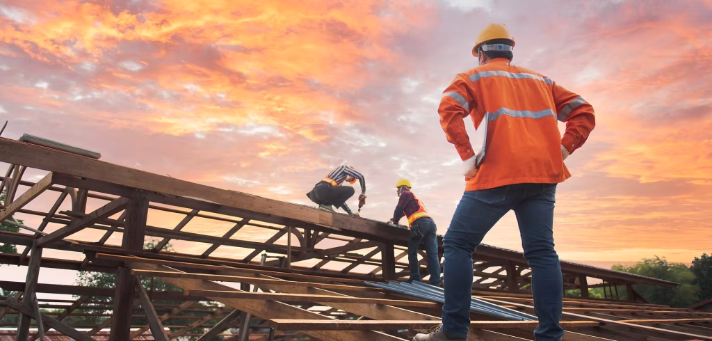 Three construction workers in safety gear and helmets work on a building’s roof structure at sunset in NY, with a dramatic orange sky behind them. One observes while two handle beams, highlighting home construction in Suffolk County.