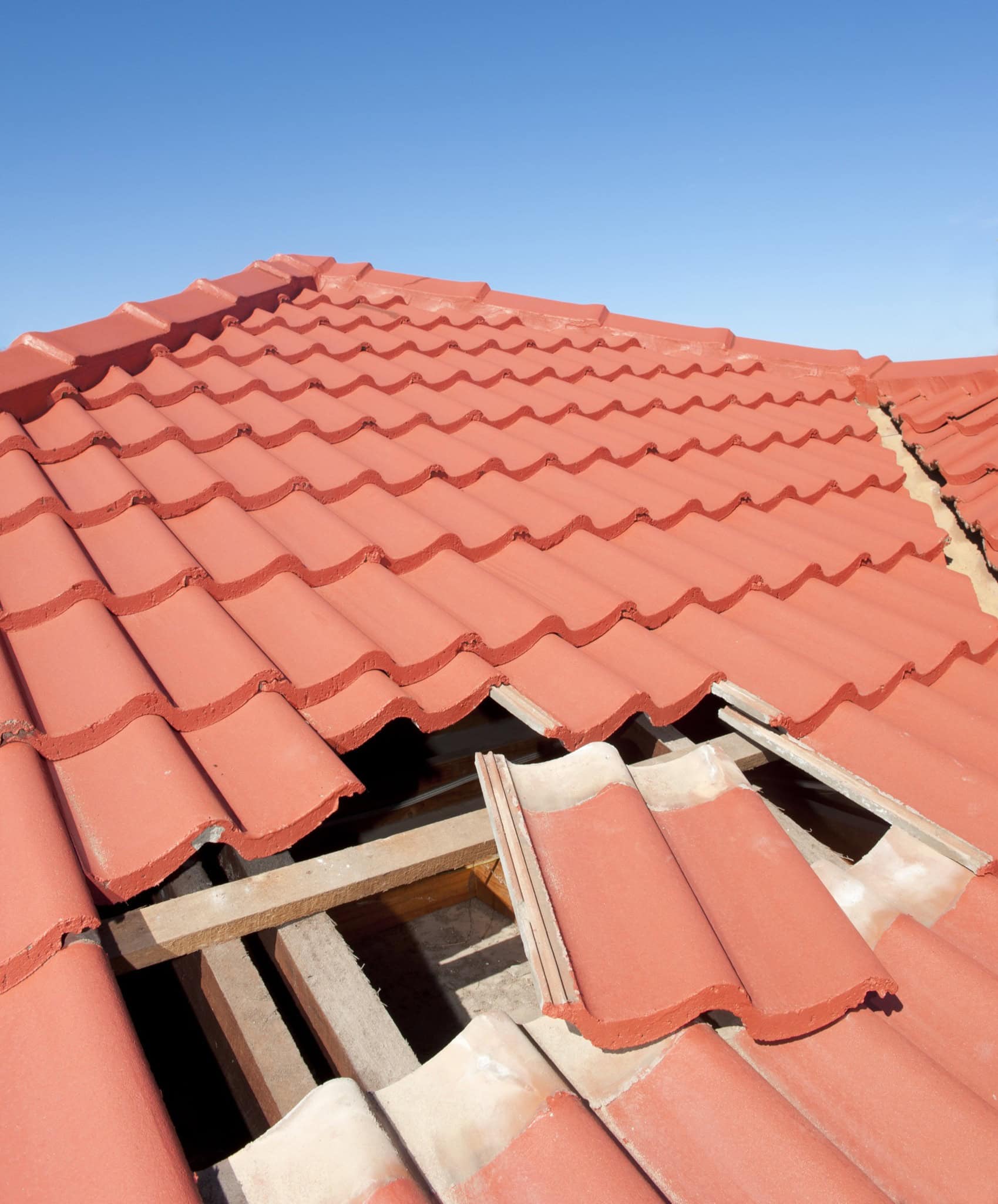 A red tiled roof with several missing and broken tiles, exposing wooden beams underneath, stands as a reminder of the importance of quality home construction in Suffolk County, NY, set against a clear blue sky.