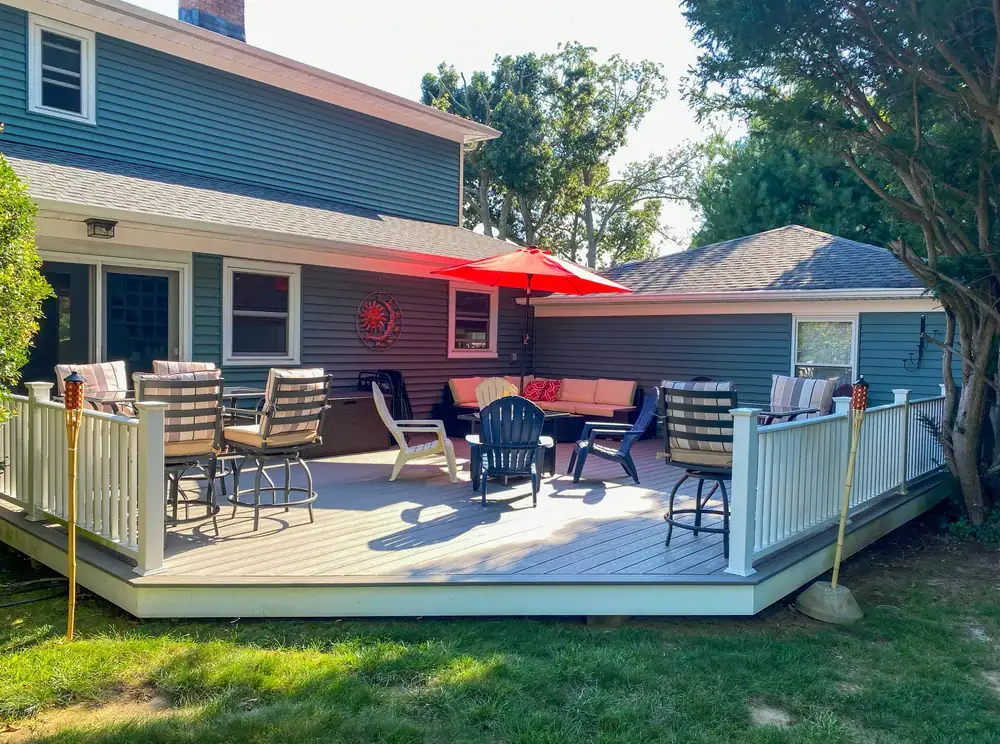 A backyard deck attached to a blue house in NY features cushioned patio chairs, a red umbrella, a sofa, a fire pit, and tiki torches. Built by home construction Suffolk County experts, the deck overlooks a grassy lawn and is bordered by a white railing.