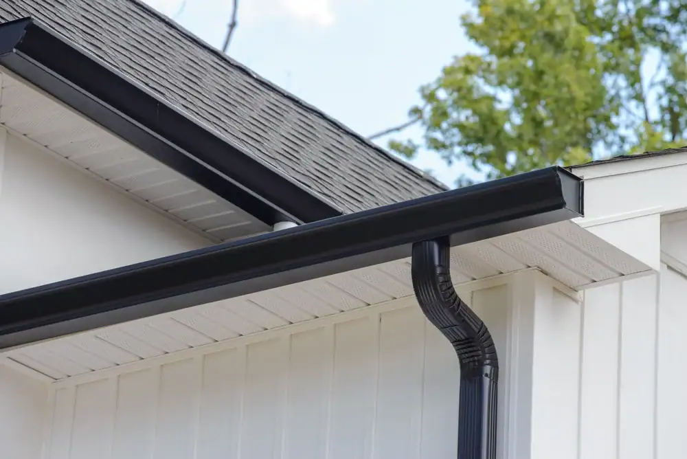 Close-up of a black rain gutter and downspout attached to the roof edge of a white house, reflecting quality home construction in Suffolk County, NY. Trees and blue sky are visible in the background.