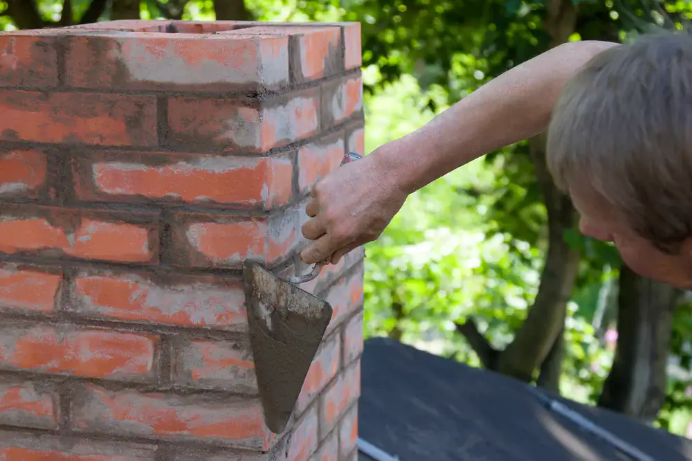 A person uses a trowel to apply mortar to a red brick chimney outdoors during a home construction project in Suffolk County, NY, with trees and greenery visible in the background.