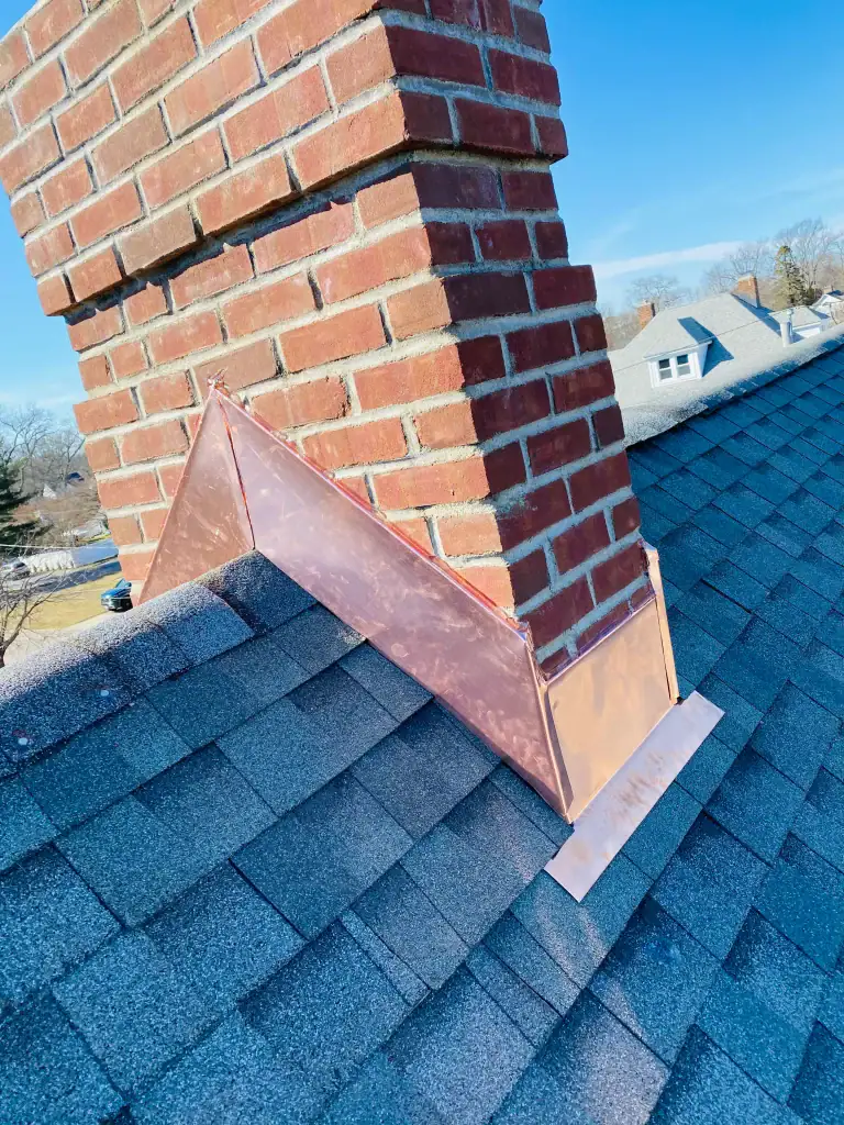 A red brick chimney with new copper flashing sits on a gray shingled roof under a clear blue NY sky, with nearby houses in Suffolk County visible in the background.