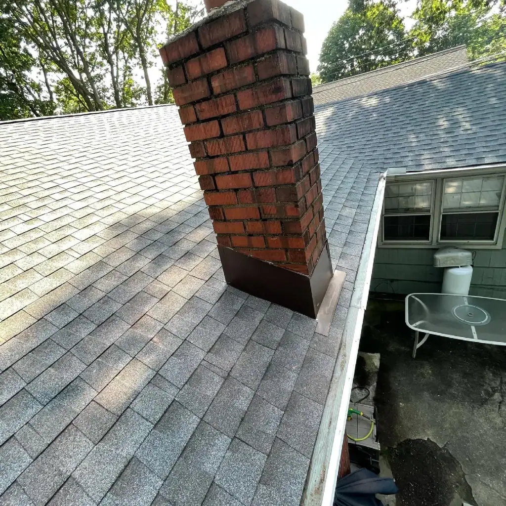 A brick chimney extends from the gray shingled roof of this NY home. Nearby is a gutter along the roof’s edge. Below, a white metal table and chairs sit on a concrete patio next to a windowed wall, with trees in the background.