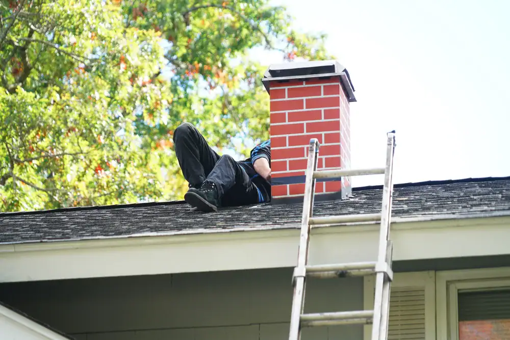 A person lies on a shingled roof next to a brick chimney, partially hidden from view—a scene common during home construction in Suffolk County, NY. A metal ladder is propped against the roof, with green trees visible in the background.