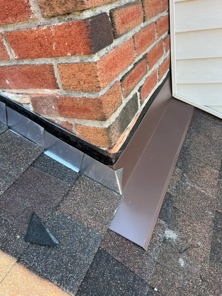 Newly installed metal flashing around the base of a brick chimney and beige siding, set over brown roof shingles, showing clean lines and precise fit—expert home construction in Suffolk County, NY to prevent water intrusion.