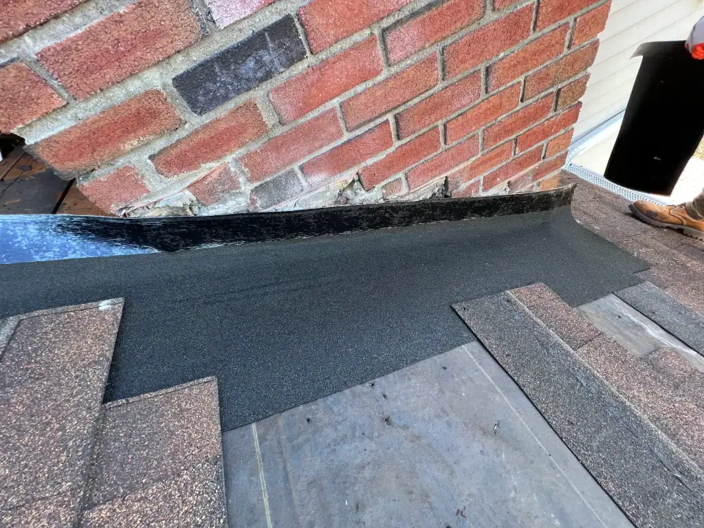 Close-up of roof flashing installation in home construction Suffolk County, NY, where black metal flashing and underlayment meet a brick chimney. Asphalt shingles are partially installed nearby, with a worker’s boot visible on the right.