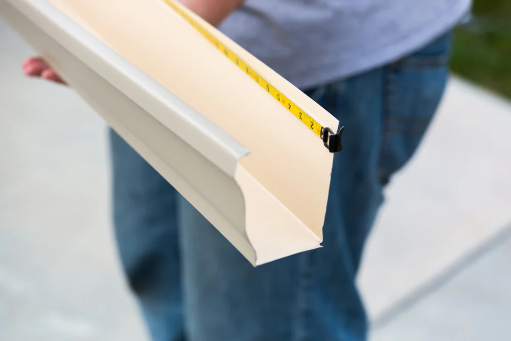 A person in jeans measures a beige rain gutter with a tape measure outdoors on a paved surface, likely part of home construction in Suffolk County, NY.