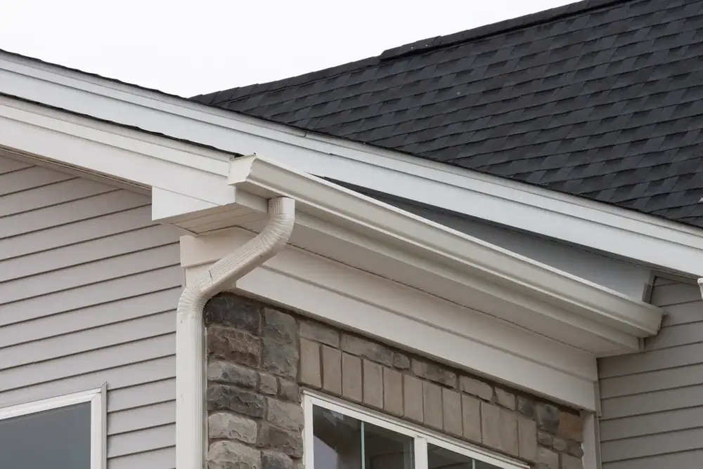 Close-up of the corner of a NY house exterior, highlighting beige siding, stonework, and a white rain gutter along the roof edge—showcasing quality home construction in Suffolk County with dark asphalt shingles above.