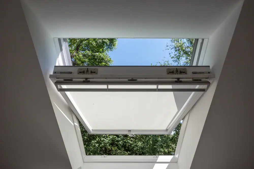 Open skylight window set in a white ceiling, with a view of green tree tops and blue sky—sunlight illuminates the frame. Perfect for brightening any space, this feature is popular in home construction across Suffolk County, NY.
