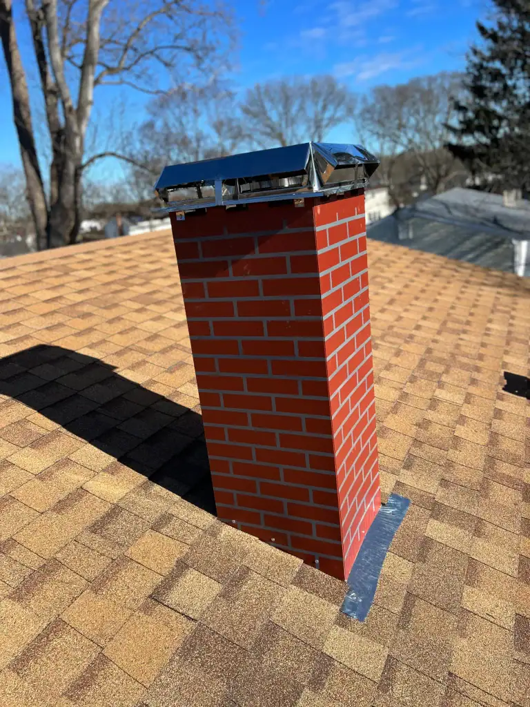 A red brick chimney with a shiny metal cap stands on a tan shingle roof under a clear NY sky, with bare trees and rooftops—hallmarks of home construction Suffolk County—in the background.