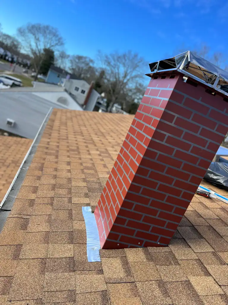 A red brick chimney with shiny metal flashing is installed on a sloped shingle roof, showcasing quality home construction in Suffolk County, NY. Suburban houses and leafless trees appear in the background under a blue sky.