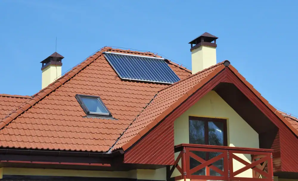 A house in NY with a red-tiled roof showcases a solar panel and skylight—ideal for modern home construction in Suffolk County. Two yellow chimneys with dark caps, a balcony with red rails, and a clear blue sky complete the scene.