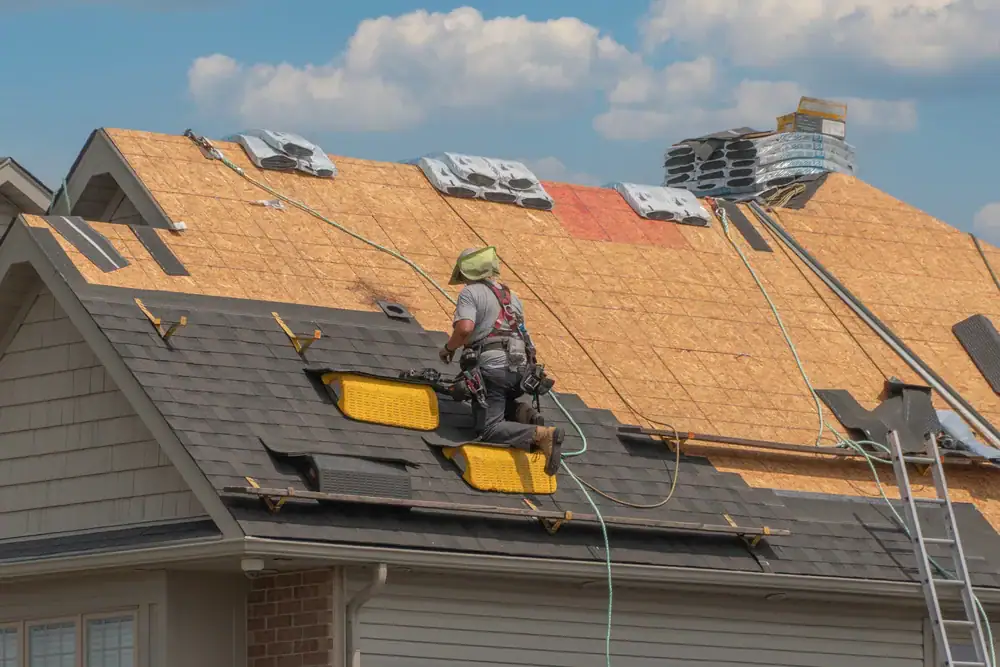 A construction worker wearing safety gear installs shingles on the roof of a house under a partly cloudy sky, with stacks of materials visible—showcasing home construction in Suffolk County, NY.