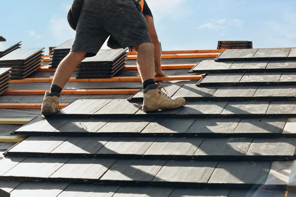 A person in work boots and shorts installs black slate tiles on a roof under a blue sky, with stacked roofing materials nearby—a typical scene in home construction projects across Suffolk County, NY.