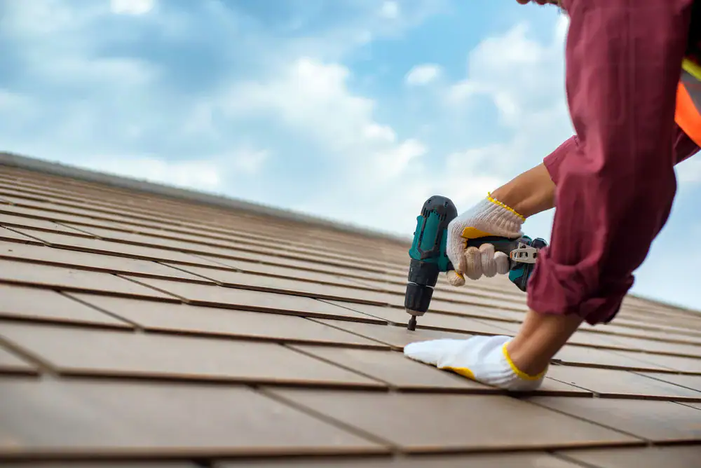 A person wearing gloves uses a power drill to fasten shingles on a rooftop in Suffolk County, NY, showcasing expert work in home construction under a partly cloudy sky.