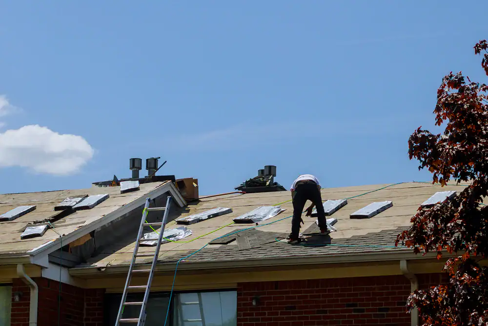 A worker is installing shingles on a house roof in Suffolk County, NY, surrounded by various roofing materials. A ladder leans against the roof, and a tree's leaves are visible on the right under a clear blue sky.