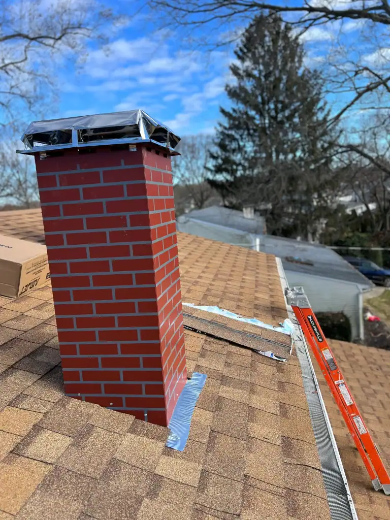 A red brick chimney with a metal cap is installed on a brown shingle roof, showcasing skilled home construction in Suffolk County, NY. Silver flashing surrounds the chimney base, with a level and tape measure nearby. Trees and houses are visible in the background.