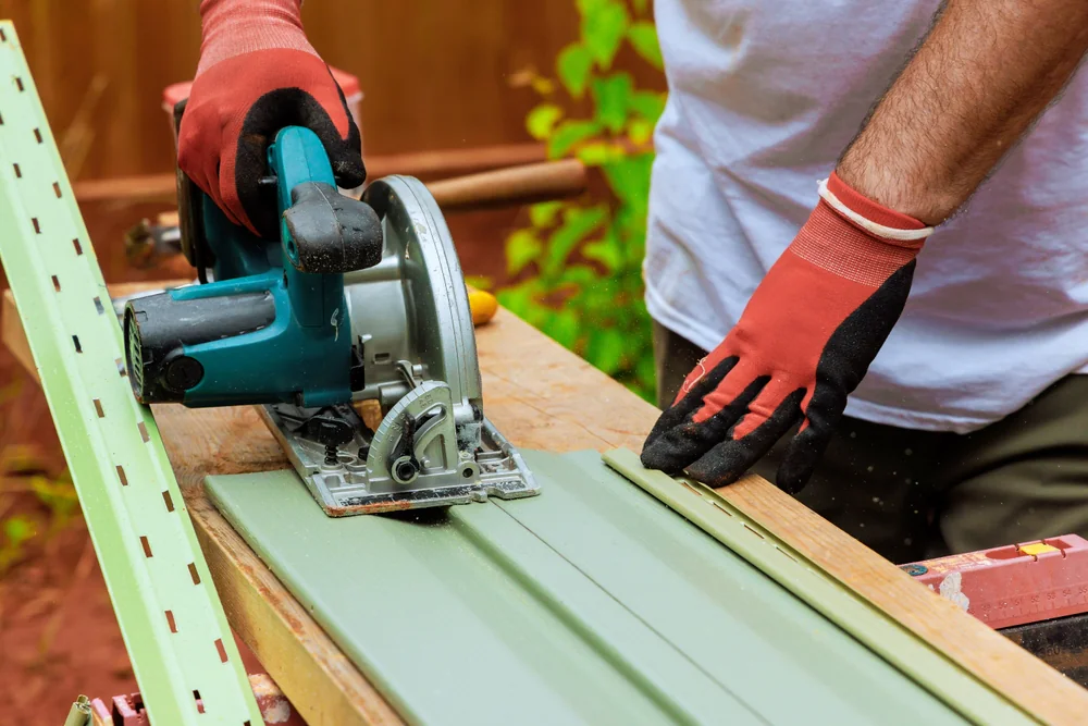 A person wearing red gloves uses a circular saw to cut a green vinyl siding panel on a wooden workbench outdoors during a home construction project in Suffolk County, NY.