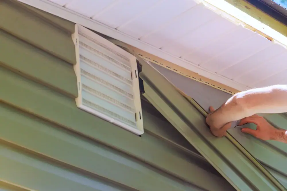 Close-up of a person’s hands installing green vinyl siding on the exterior wall of a house in Suffolk County, NY, near a white vent and under the roof eaves—capturing a real moment in home construction.