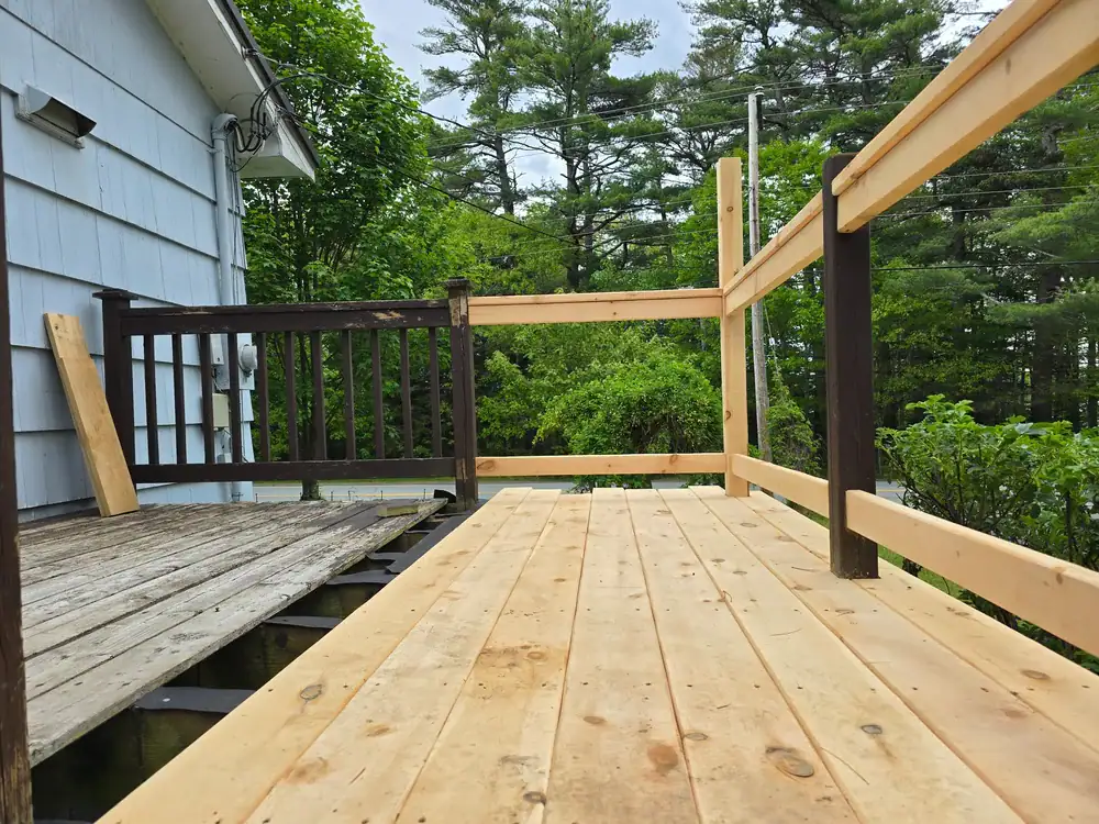 A wooden deck under construction outside a house in Suffolk County, NY, with new light-colored boards and partially built railing beside an older, weathered deck, all surrounded by trees and greenery.