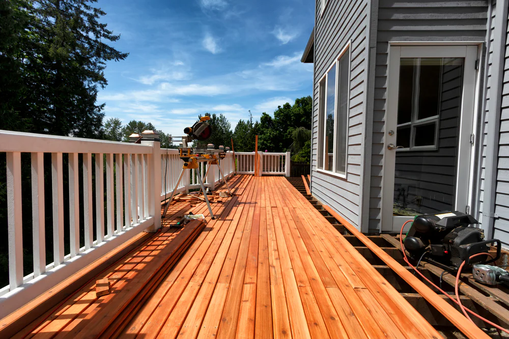 A freshly built wooden deck attached to a gray house in Suffolk County, NY, with construction tools and equipment present, surrounded by white railings and trees under a blue sky.