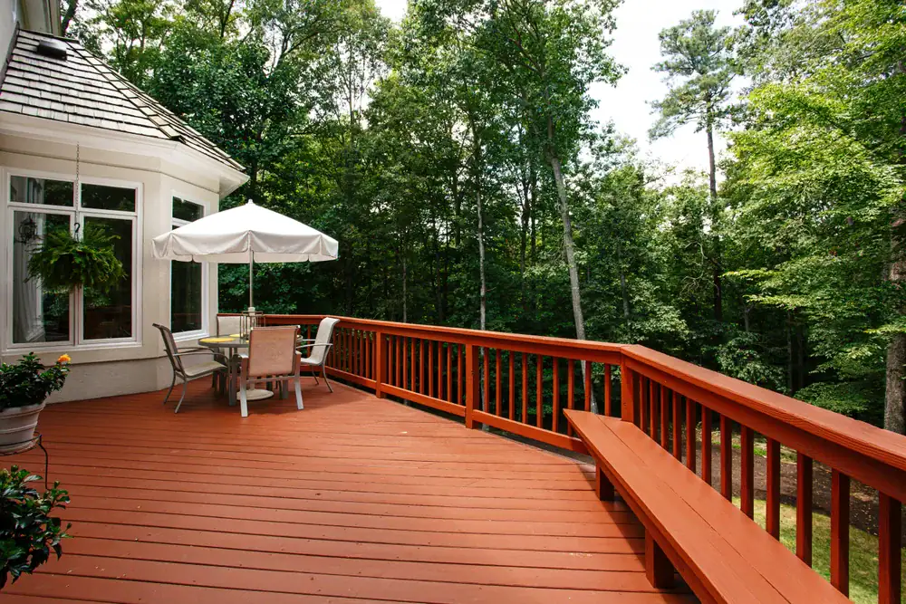 A spacious wooden deck with red-brown flooring, a built-in bench, patio table, chairs, and a white umbrella—crafted by experts in home construction in Suffolk County, NY—surrounded by lush trees and attached to a house with large windows.