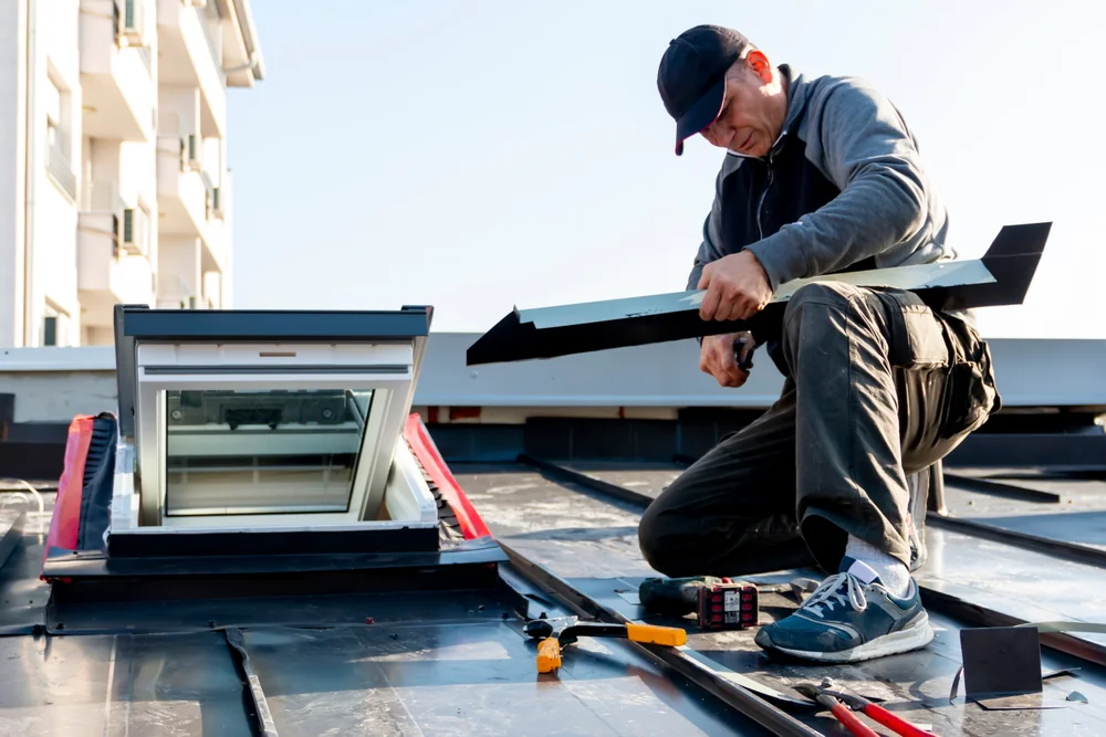 A person kneels on a NY rooftop, installing or repairing a skylight window. Tools and materials are spread around, and the person is holding a metal frame piece while wearing casual work clothes and a cap—typical of home construction Suffolk County.