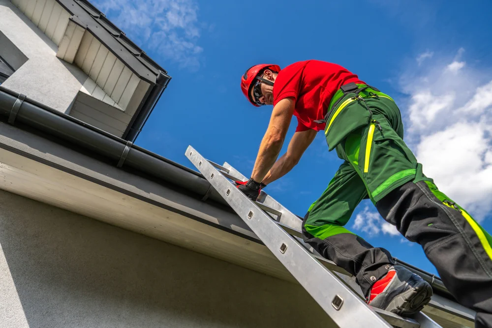 A worker wearing safety gear stands on a ladder, inspecting or repairing the gutter of a modern house under a clear blue sky—a common scene in home construction Suffolk County, NY.