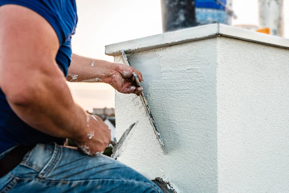A person in a blue shirt applies white plaster to a building’s exterior wall in Suffolk County, NY, using a trowel, with paint splatters visible on their arm and clothes—a typical scene in home construction.