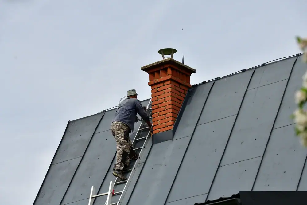 A person wearing a hat and camouflage pants climbs a ladder leaned against a steep metal roof, working near a red brick chimney under a cloudy sky—a typical scene in NY home construction Suffolk County.