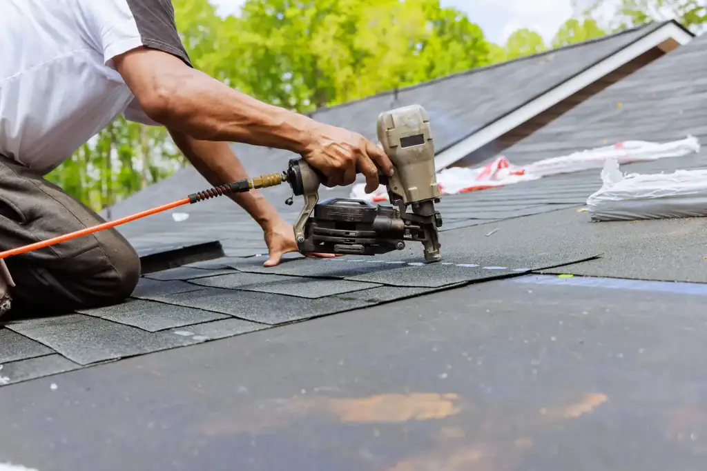 A person uses a nail gun to install asphalt shingles on a house roof in Suffolk County, NY, surrounded by trees. Roofing materials and tools are scattered nearby.