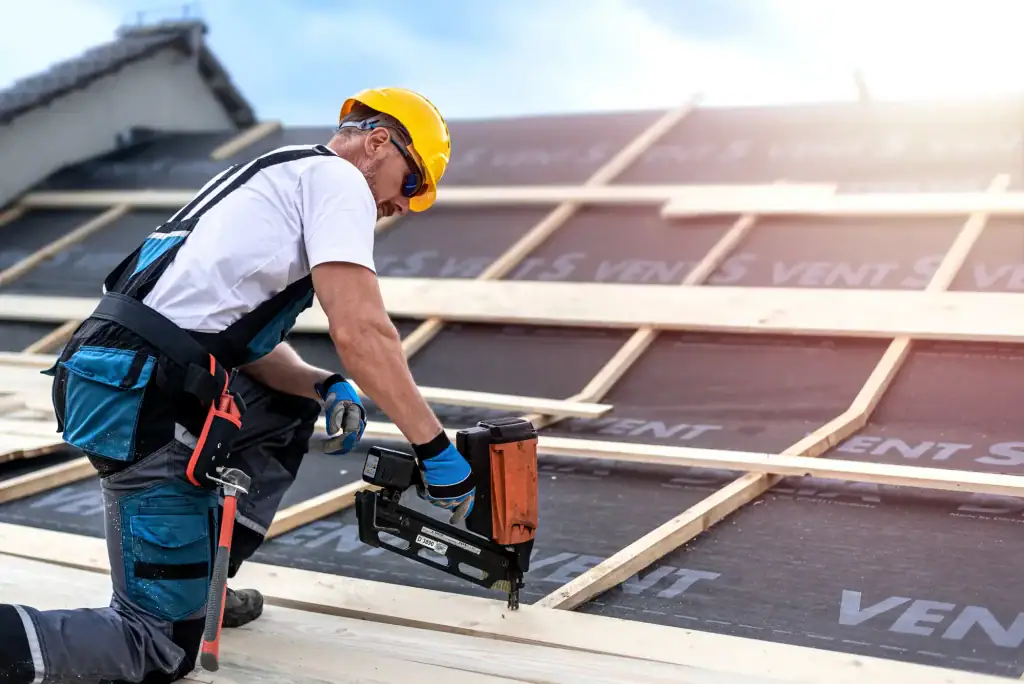 A construction worker in a yellow hard hat and gloves uses a nail gun to secure wooden beams on a roof structure under bright daylight during a Home Construction Suffolk County, NY project.