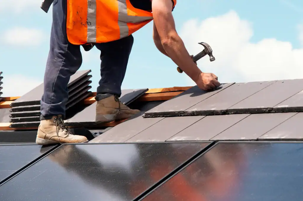 A construction worker in an orange safety vest installs roof tiles with a hammer next to solar panels on a rooftop under a blue sky, showcasing Home Construction Suffolk County, NY.