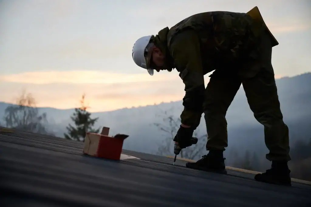 A construction worker wearing a white hard hat and camouflage gear uses a power drill on a rooftop during sunset in Suffolk County, NY.