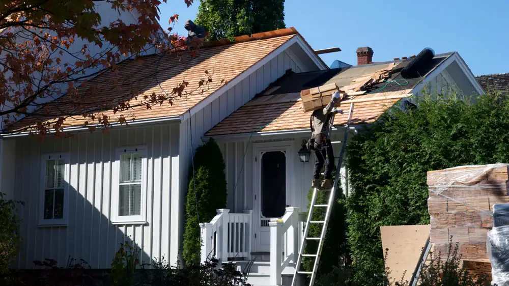 A worker climbs a ladder carrying roofing materials onto the roof of a white house, where new shingles are being installed. Construction materials are stacked nearby and trees surround the house.