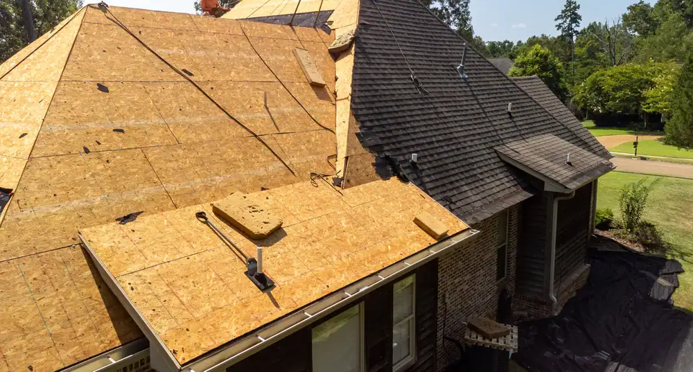 A house with part of its roof under construction, showing exposed wooden sheathing and some installed shingles. Roofing tools and materials are visible, and green trees surround the property.