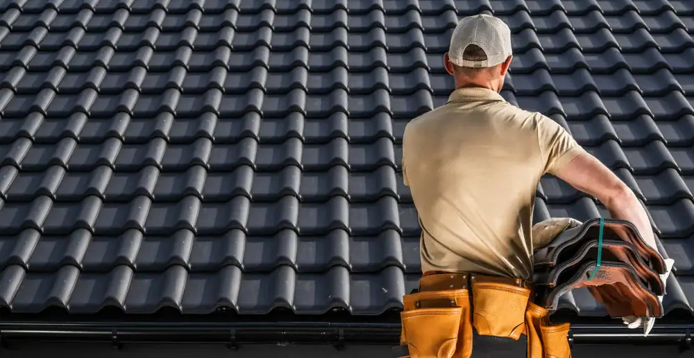 A roofer wearing a cap and tool belt stands on a roof, holding a bundle of roof tiles with dark, wavy shingles laid out across the roof in front of him.
