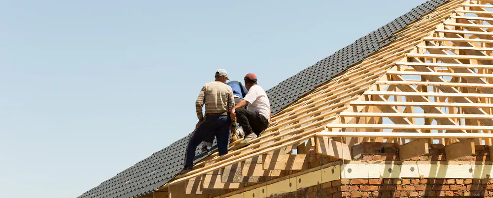Three construction workers sit on the wooden frame of a house roof under construction, with a clear blue sky in the background.